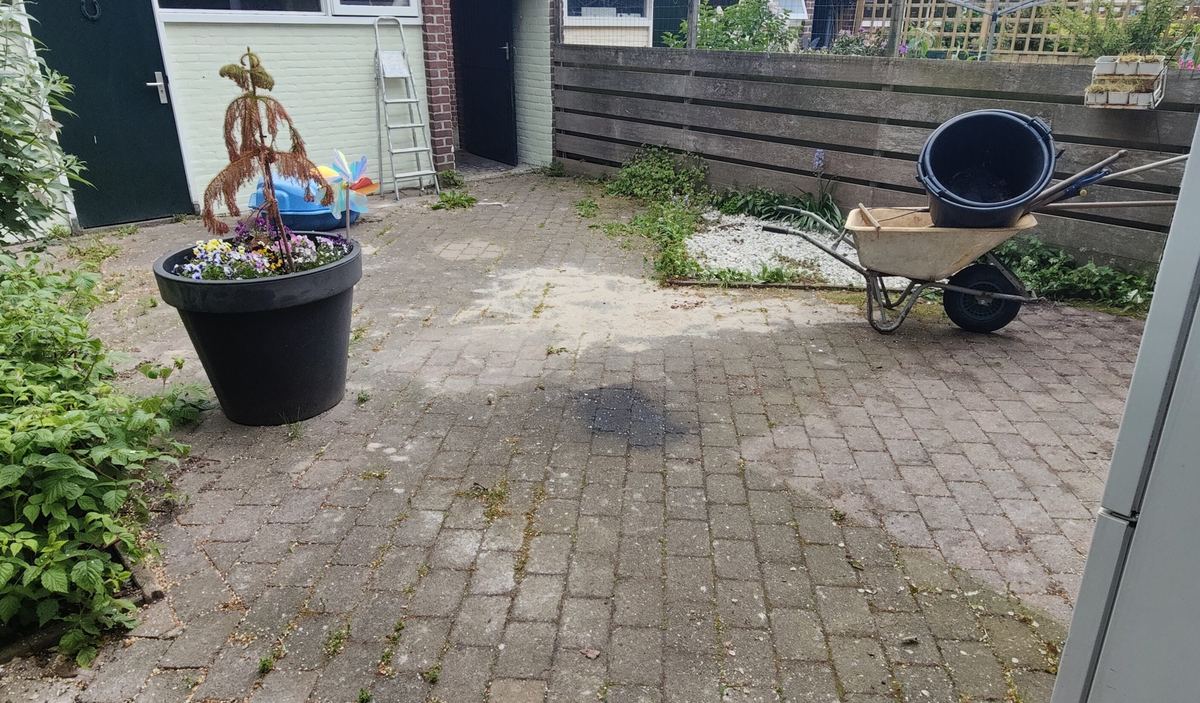 Terras van oude klinkers met onkruid in de voegen, een plantenbak en een kruiwagen